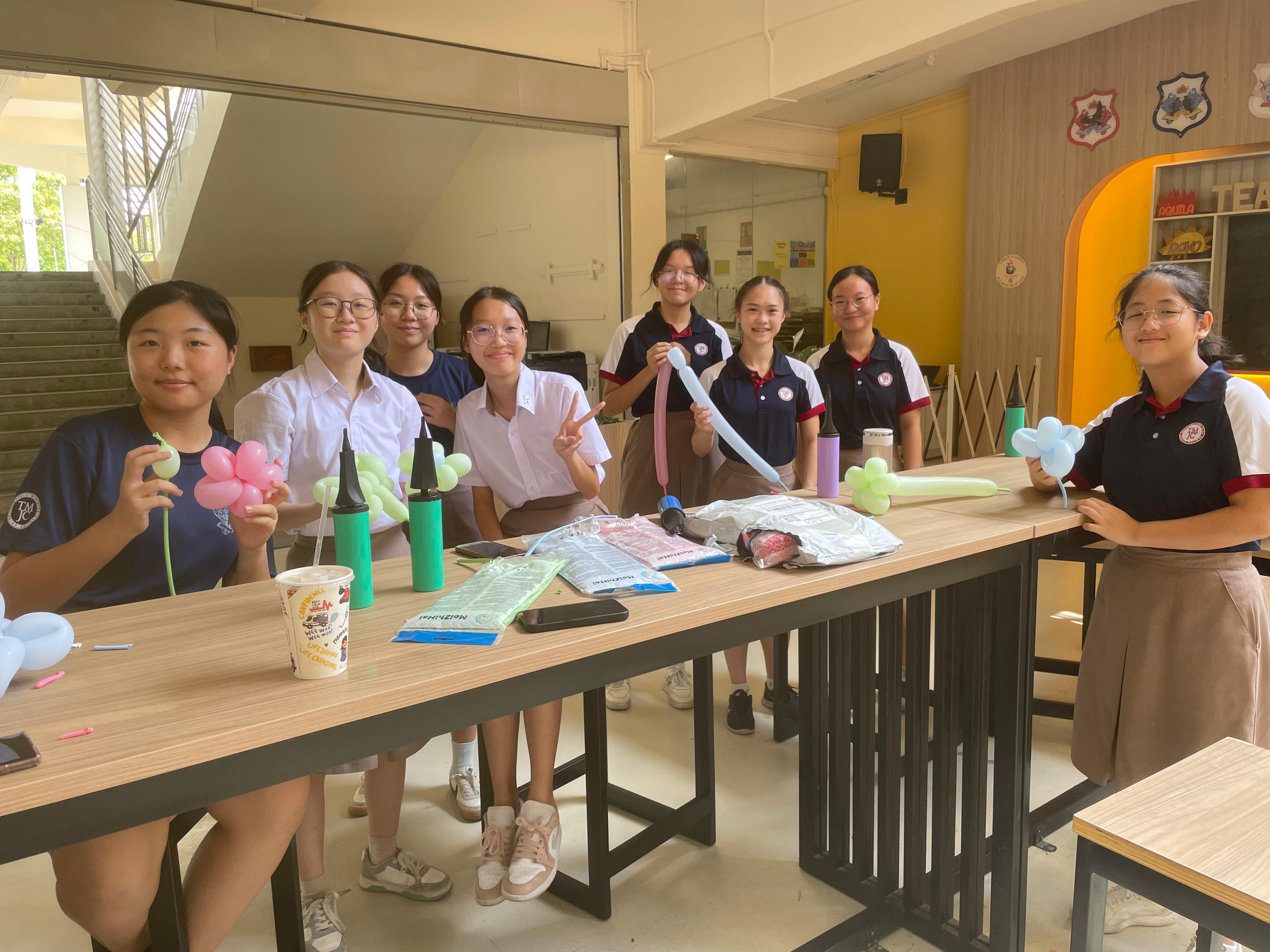 Enter a descriptiveEight female students in school uniforms smile and pose together at a long table in a casual indoor common area. Several of them hold up small balloon flowers and balloon sculptures they have crafted. The table is scattered with balloon-making supplies, including pump inflators and packets of balloons in pastel colours. alt text.